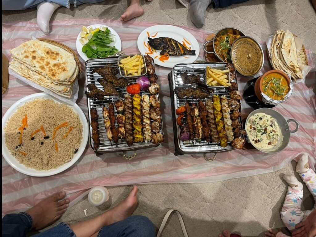  Overhead view of a lavish Middle Eastern feast spread out on a plastic mat on the floor. The spread includes two large grills filled with various meat skewers and french fries, a large platter of rice with carrots and raisins, flatbread, grilled fish, stews, and side dishes, with family members gathered around.
