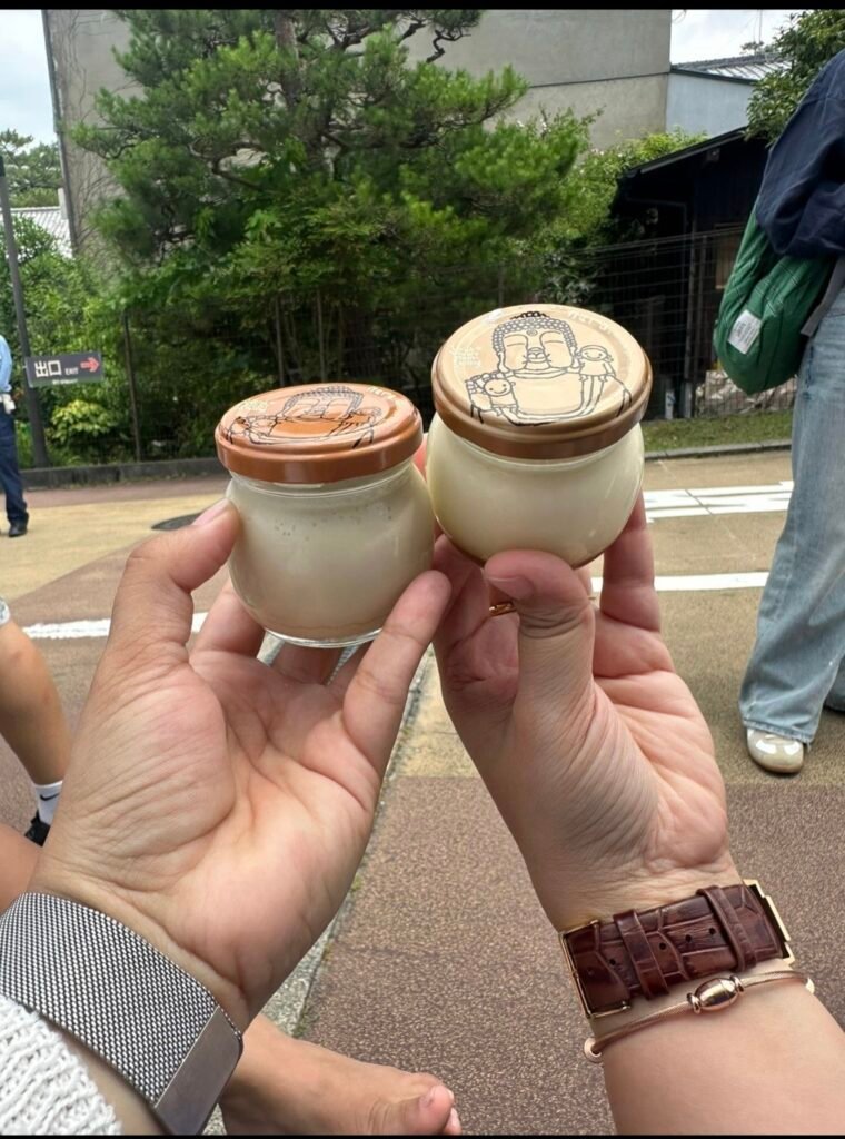 Two small glass jars of Mahoroba Daibutsu pudding with printed Buddha lids held up outdoors in Nara.