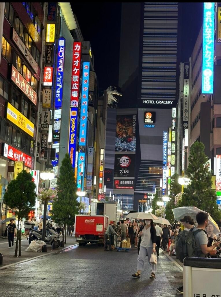 `Illuminated night street in Shinjuku lined with neon signs, leading toward the giant Godzilla head atop Hotel Gracery.`
