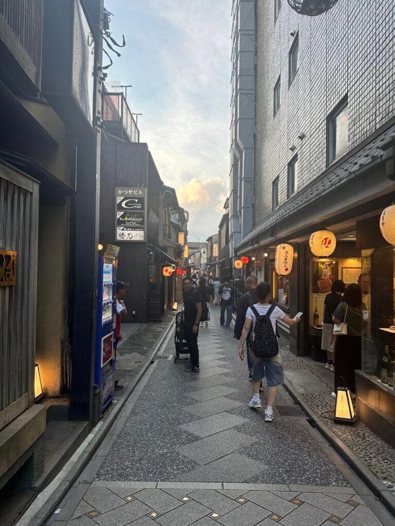 A narrow stone-paved pedestrian alley lined with traditional storefronts, glowing paper lanterns, and visitors walking in Pontocho Alley, Kyoto.

