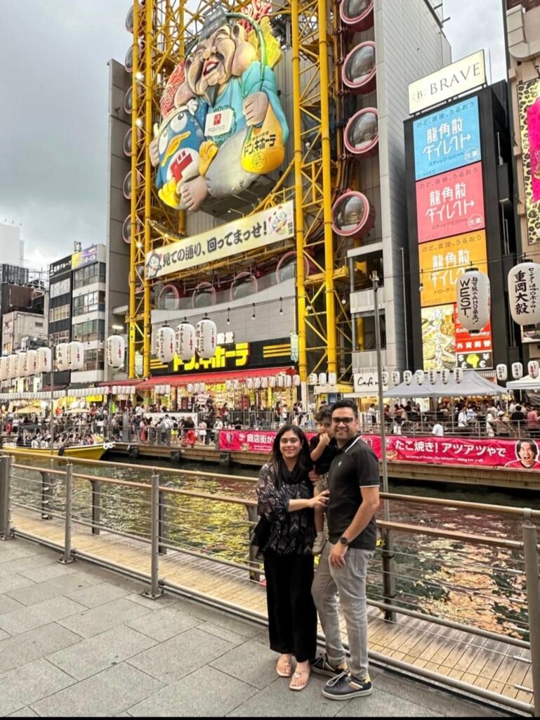 family posing along dotonbori canal walkway in front of yellow don quijote ferris wheel building in osaka 