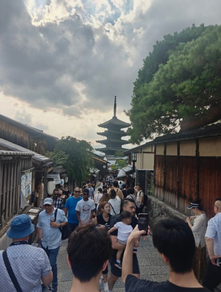 Tourists walking down the historic sloped street of Ninenzaka with traditional wooden Machiya buildings and the Yasaka Pagoda rising in the background in Kyoto.