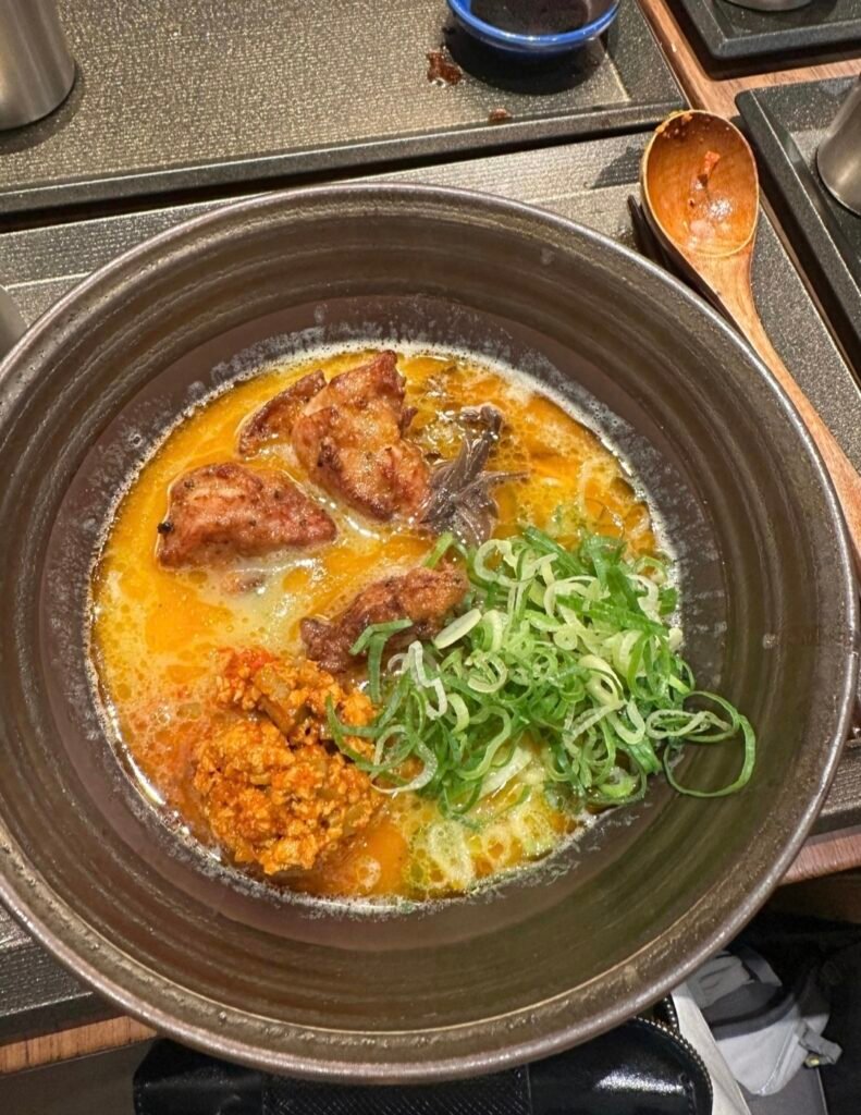 A rich bowl of Japanese ramen topped with crispy chicken pieces, green onions, and chili paste in a dark ceramic bowl AT Honolu Shinjukugyoenmae Halal Ramen 