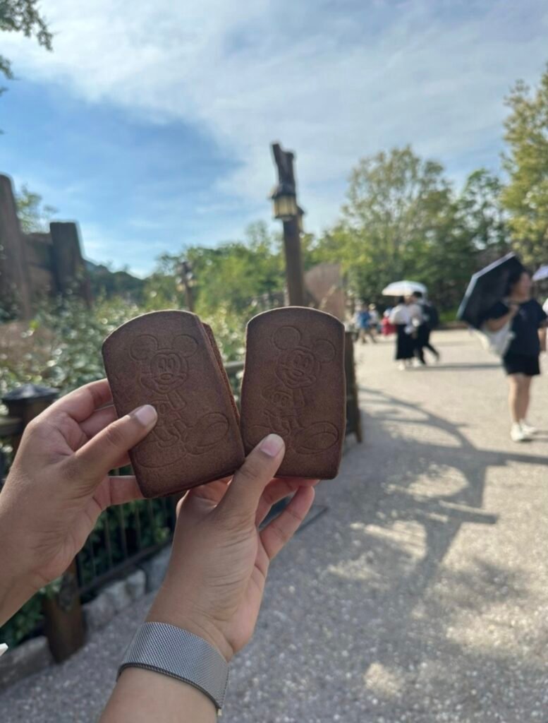 Two hands holding Mickey Mouse ice cream sandwiches outdoors at Tokyo DisneySea with walkways and trees in the background.`