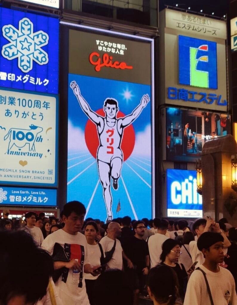 THE ICONIC GLICO MAN SIGN GLOWING OVER THE BUSTLING EVENING CROWD AT EBISU BRIDGE IN DOTONBORI , OSAKA 
