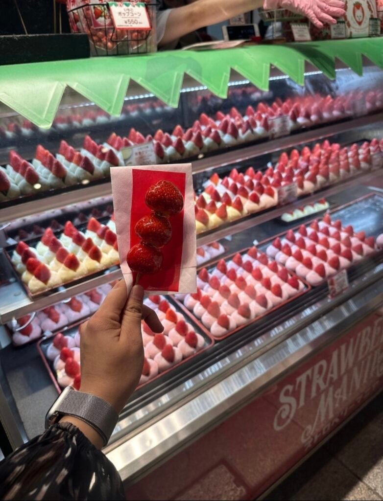 A SKEWER OF GLOSSY CANDIED STRAWBERRIES HELD IN FRONT OF A STRAWBERRY MANIA DESSERT GLASS DISPLAY CASE IN OSAKA 