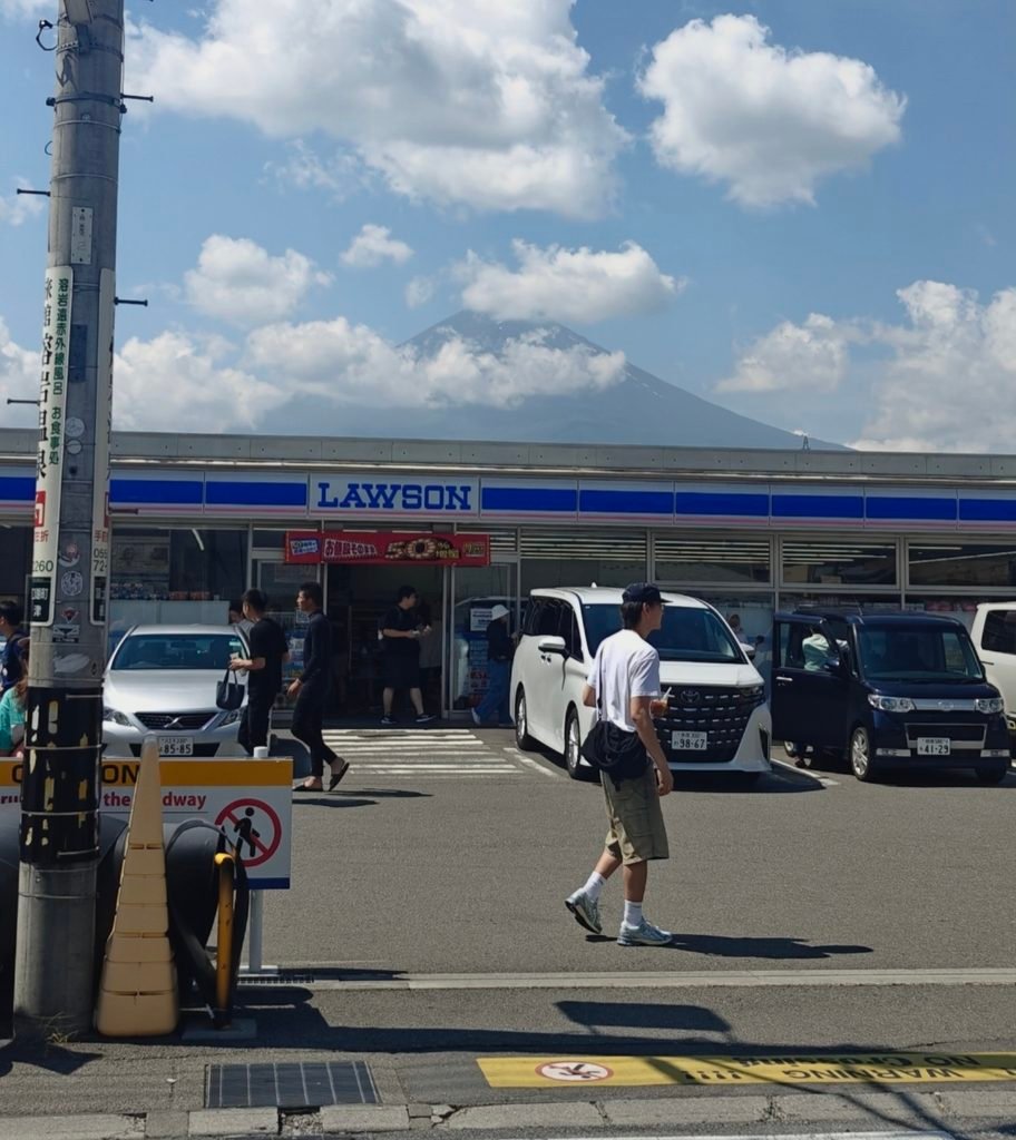 `A clear view of a Lawson convenience store parking lot with Mount Fuji towering in the background under a sunny blue sky.`