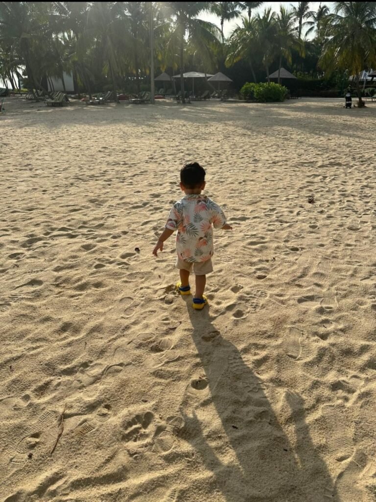 Young toddler in a printed short-sleeve shirt walking on a sandy tropical beach lined with palm trees.