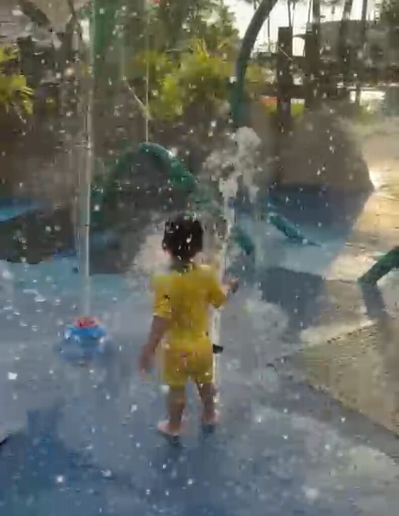 Toddler playing in a outdoor water spray splash pad on a sunny day.