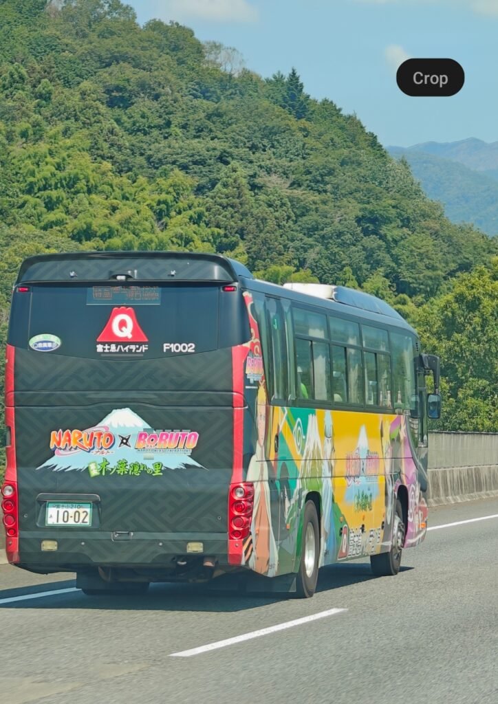 A tour bus featuring full-body Naruto and Boruto anime wrap graphics driving along a scenic highway surrounded by lush hills.`
