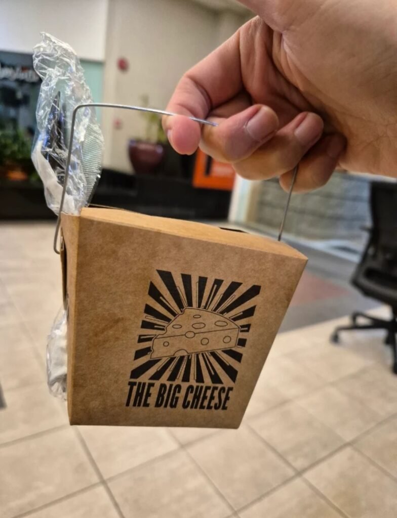 Hand holding a takeaway paper box labeled "The Big Cheese" featuring a cheese wedge illustration.