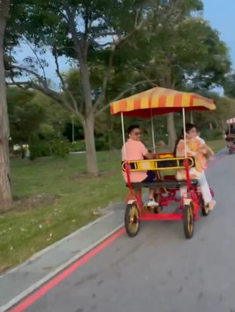 Family riding a red and yellow quadricycle surrey bike along a paved park path.