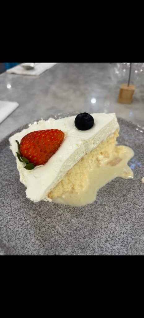 A delicious slice of white tres leches milk cake topped with a fresh strawberry and blueberry on a grey plate.