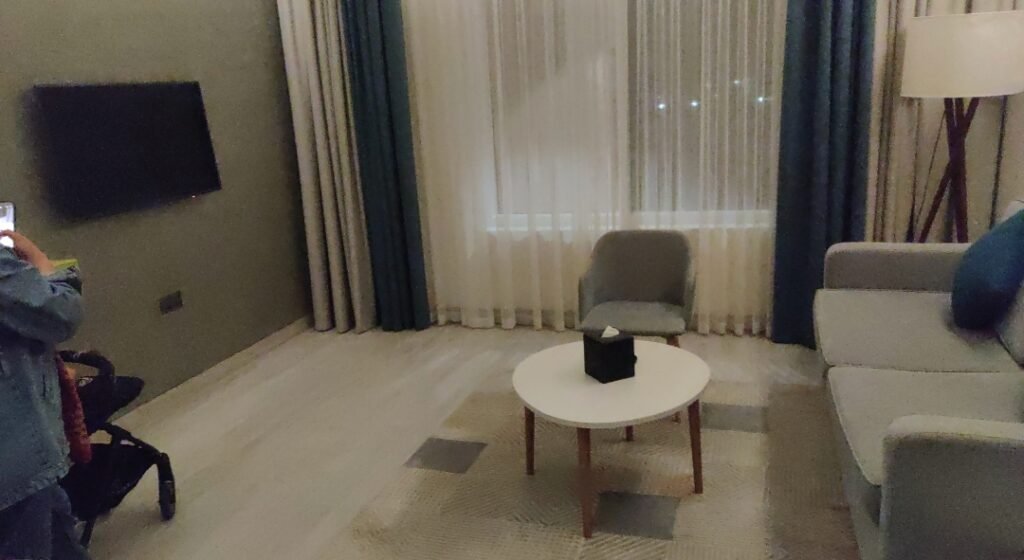 A cozy hotel suite living room setup with a grey sofa, round coffee table, flat screen TV, and curtains.