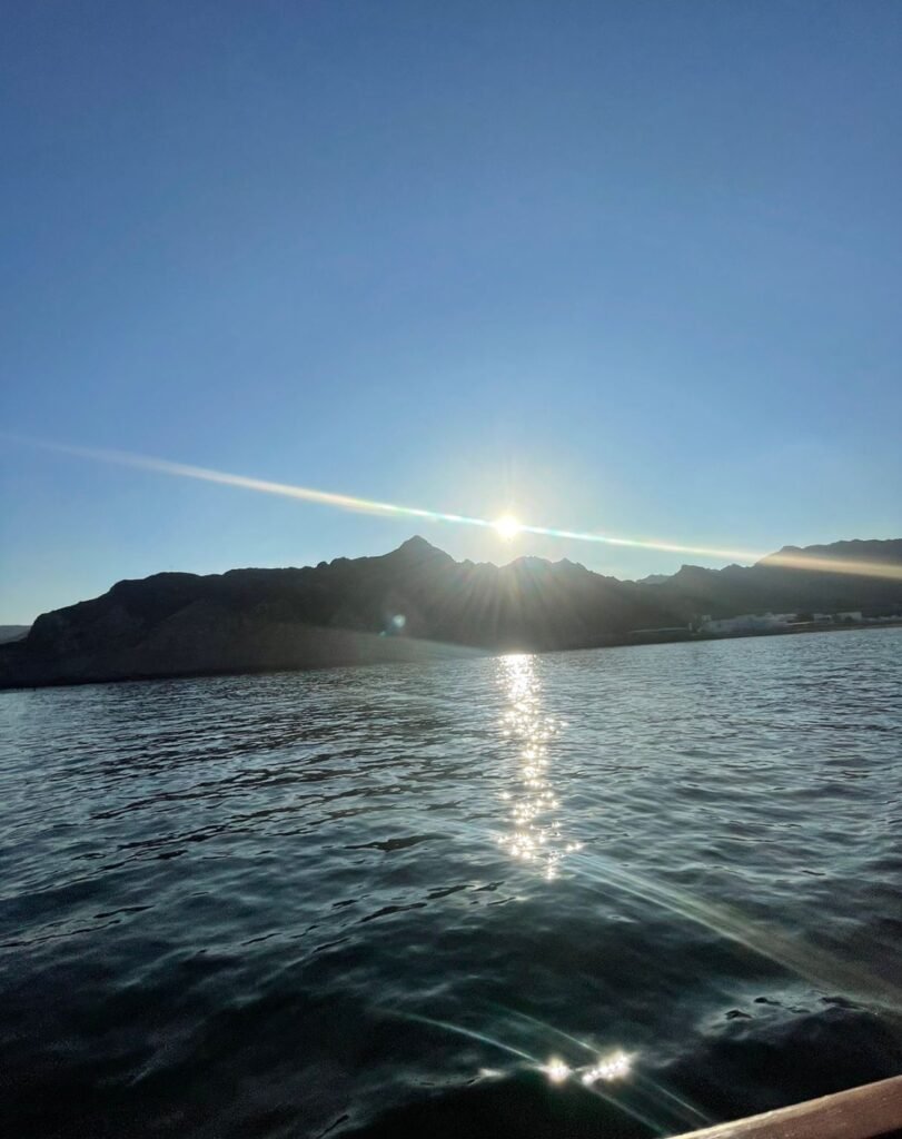 Alt Text: Scenic view of the sun setting over the coastal mountains of Muscat, Oman, with sunbeams reflecting across the ocean water.